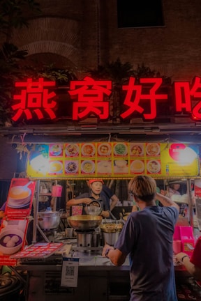 A bustling street food stall is illuminated with vibrant signage in red Chinese characters. The vendor, wearing a cap, is preparing food, surrounded by pots and cooking utensils. A customer stands in front, seemingly placing an order. Menu items are displayed with pictures above the stall.