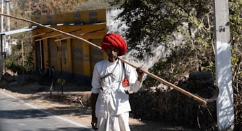 A person wearing traditional attire and a vibrant red turban holds a long, wooden stick over their shoulder. They stand by a roadside with a yellow building and trees in the background.
