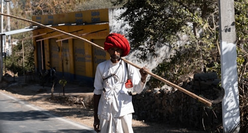 A person wearing traditional attire and a vibrant red turban holds a long, wooden stick over their shoulder. They stand by a roadside with a yellow building and trees in the background.