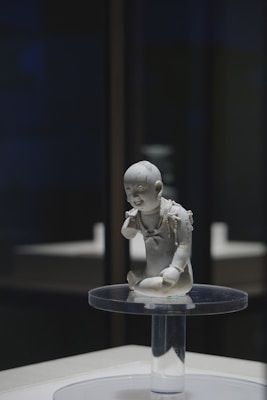 A small, detailed sculpture of a child is displayed on a clear glass pedestal. The child is depicted in a kneeling pose, adorned with intricate jewelry including necklaces and armlets.
