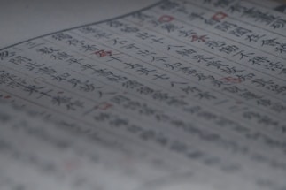 A close-up of a page containing text written in both blue and red ink. The characters appear to be of East Asian origin, possibly Japanese or Chinese. The focus is on the central area of the page, with some blurring at the edges. Red ink highlights certain words or symbols throughout the text.