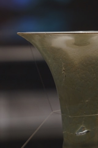 A close-up view of a ceramic object with a smooth, curved edge and a greenish glazed surface. The glaze is slightly crackled, suggesting age or intentional design. The lighting creates subtle reflections on the surface, highlighting the texture and color variations.