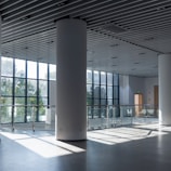 A spacious and modern interior with large floor-to-ceiling windows allowing natural light to stream in, illuminating the polished floor. The area features white cylindrical columns and a glass railing on an upper level. The ceiling has a grid pattern with integrated lighting fixtures.