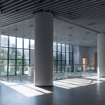 A spacious and modern interior with large floor-to-ceiling windows allowing natural light to stream in, illuminating the polished floor. The area features white cylindrical columns and a glass railing on an upper level. The ceiling has a grid pattern with integrated lighting fixtures.