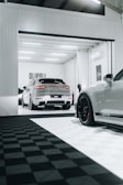 A modern garage space with sleek, luxury cars. One car is parked inside the garage, while another is partially visible in the foreground, showcasing its stylish rims. The area is well-lit, featuring a clean, minimalistic design with a predominantly white and black color scheme.