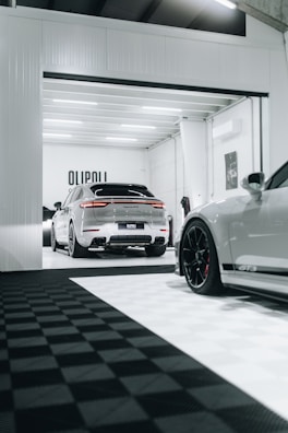 A modern garage space with sleek, luxury cars. One car is parked inside the garage, while another is partially visible in the foreground, showcasing its stylish rims. The area is well-lit, featuring a clean, minimalistic design with a predominantly white and black color scheme.