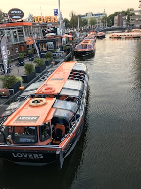 Several canal tour boats with orange roofs are docked along a busy waterfront. The area features a modern cafe with large windows and signage. A row of potted plants lines the dockside, while people can be seen inside the boats and walking by the water. Buildings and a bridge span the background, creating a bustling urban scene.