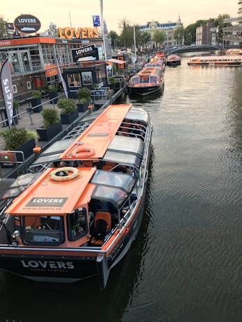 Several canal tour boats with orange roofs are docked along a busy waterfront. The area features a modern cafe with large windows and signage. A row of potted plants lines the dockside, while people can be seen inside the boats and walking by the water. Buildings and a bridge span the background, creating a bustling urban scene.