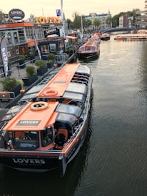 Several canal tour boats with orange roofs are docked along a busy waterfront. The area features a modern cafe with large windows and signage. A row of potted plants lines the dockside, while people can be seen inside the boats and walking by the water. Buildings and a bridge span the background, creating a bustling urban scene.