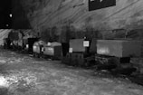 A series of mining carts line up in an underground cavern with rough, textured walls. The carts appear slightly weathered and are situated on icy or snowy ground. Some informational signs are attached to them.