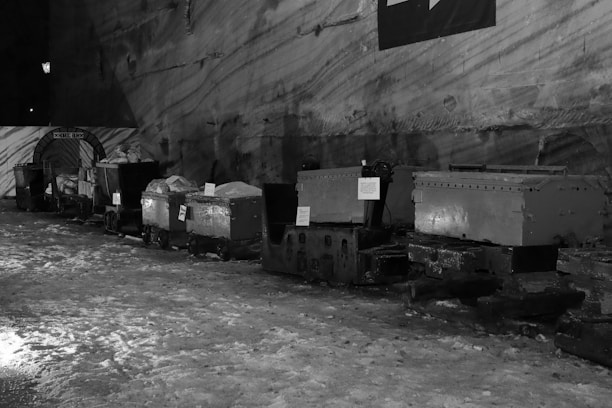 A series of mining carts line up in an underground cavern with rough, textured walls. The carts appear slightly weathered and are situated on icy or snowy ground. Some informational signs are attached to them.