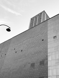A modern architectural scene with a tall building in the background, featuring numerous windows and a complex design. The foreground shows the side of another structure with a large wall made of square tiles. A streetlamp is visible in the upper left corner, adding an urban element to the composition.