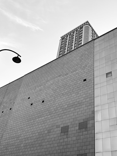 A modern architectural scene with a tall building in the background, featuring numerous windows and a complex design. The foreground shows the side of another structure with a large wall made of square tiles. A streetlamp is visible in the upper left corner, adding an urban element to the composition.
