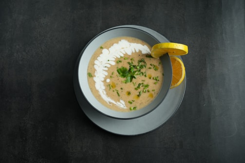 A bowl of creamy soup garnished with a swirl of cream and chopped herbs sits on a table. A lemon slice is placed on the rim of the bowl, and the soup is served in a light gray dish on a dark surface.