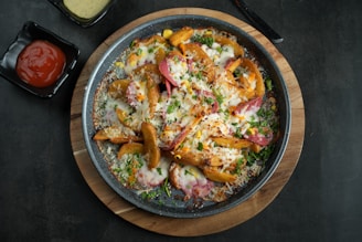 A plate filled with baked potato wedges topped with melted cheese, sliced sausages, corn, and sprinkled with chopped herbs. The plate is placed on a wooden cutting board, and there are two square sauce dishes nearby, one containing ketchup and the other containing a creamy yellow sauce.