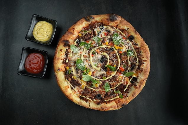 A rustic pizza topped with various ingredients, including olives, green herbs, and slices of colorful vegetables. The pizza crust is golden brown and slightly crispy, surrounded by two square black dishes containing a creamy yellow sauce and red ketchup.