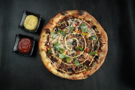 A rustic pizza topped with various ingredients, including olives, green herbs, and slices of colorful vegetables. The pizza crust is golden brown and slightly crispy, surrounded by two square black dishes containing a creamy yellow sauce and red ketchup.