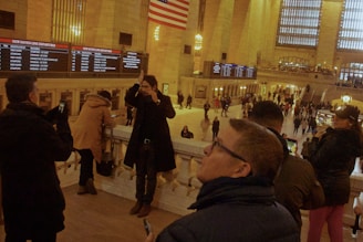 A bustling train station with people taking photos and looking at the departure board. The interior is grand, with tall columns and large windows. An American flag is visible overhead, and there are multiple levels with people observing the scene from different vantage points.