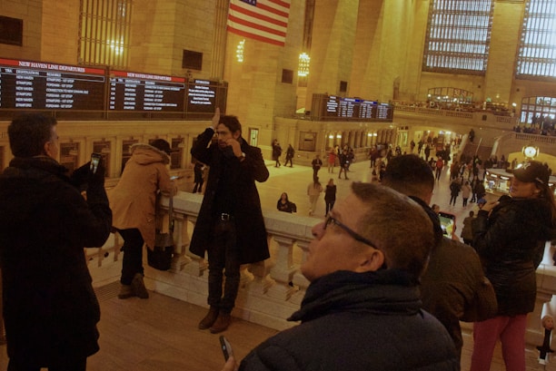 A bustling train station with people taking photos and looking at the departure board. The interior is grand, with tall columns and large windows. An American flag is visible overhead, and there are multiple levels with people observing the scene from different vantage points.