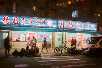 A street view of a meat market at night, with several people walking on the sidewalk in front of the store. The signage is in both Chinese and English, with the name '57 Bayard Meat Market Inc.' The store has bright lights illuminating the interior, displaying various meats. There are bicycles parked in front and bags of garbage lined up on the street. A white delivery van is parked to the right side of the image.