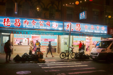 A street view of a meat market at night, with several people walking on the sidewalk in front of the store. The signage is in both Chinese and English, with the name '57 Bayard Meat Market Inc.' The store has bright lights illuminating the interior, displaying various meats. There are bicycles parked in front and bags of garbage lined up on the street. A white delivery van is parked to the right side of the image.