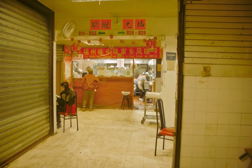 Interior of a food stall with a red sign above the entrance displaying Chinese characters and the words 'cashier' and menu numbers. A person stands inside near the counter, while another sits on a chair outside the stall. The surrounding area has closed shutters, tiled walls, and chairs with red and black seats.