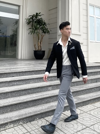 A person dressed in formal attire walks confidently on a tiled pathway. The individual is wearing a black blazer, white shirt, gray trousers, and black shoes. A potted plant is visible in the background, placed near the entrance of a modern building with large windows.