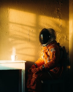 A person in an orange astronaut suit is seated in a dimly lit room next to a small white table. A steaming coffee cup is placed on the table. The room has a rough, textured wall with a warm, golden light casting shadows.