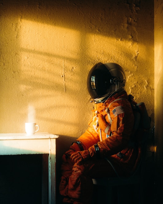 A person in an orange astronaut suit is seated in a dimly lit room next to a small white table. A steaming coffee cup is placed on the table. The room has a rough, textured wall with a warm, golden light casting shadows.