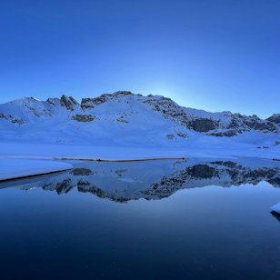 A pristine winter landscape with snow-covered mountains reflected in a calm, dark-blue lake. The sky is clear with a soft gradient from blue to white, suggesting an early morning or late afternoon setting.