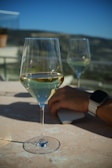 A close-up shot of a wine glass filled with white wine placed on a marble surface. In the background, another wine glass is partially visible. A hand with a wristwatch and smartphone rests on the table. The scene takes place outdoors with blurred hills and a clear blue sky.