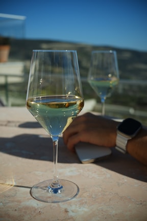 A close-up shot of a wine glass filled with white wine placed on a marble surface. In the background, another wine glass is partially visible. A hand with a wristwatch and smartphone rests on the table. The scene takes place outdoors with blurred hills and a clear blue sky.
