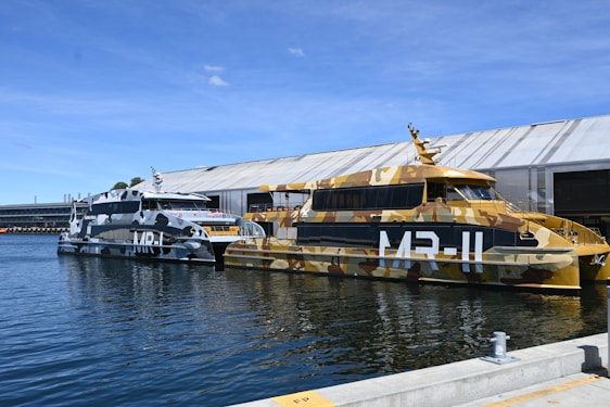 Two boats with military-style camouflage paint are docked in a waterway. One boat features a blue, gray, and white color scheme, while the other has a brown, beige, and green pattern. Both are positioned next to a large building structure.