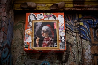 A wall adorned with various graffiti and stickers, featuring a central framed artwork of a person wearing sunglasses, a Santa hat, and a festive sweater, holding a cigarette. Above the artwork, a pair of old shoes is placed.