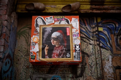 A wall adorned with various graffiti and stickers, featuring a central framed artwork of a person wearing sunglasses, a Santa hat, and a festive sweater, holding a cigarette. Above the artwork, a pair of old shoes is placed.
