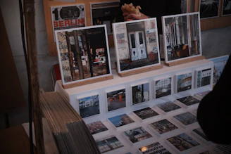 A display of art prints and photographs featuring urban and graffiti-covered scenes is set up on a table. Larger framed images rest on wooden stands, while a series of smaller prints are laid out before them. There are also visible stacks of prints to the side. Behind the display, a person holding a sandwich is partially visible.