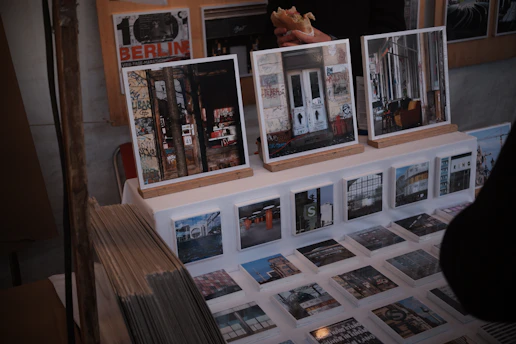 A display of art prints and photographs featuring urban and graffiti-covered scenes is set up on a table. Larger framed images rest on wooden stands, while a series of smaller prints are laid out before them. There are also visible stacks of prints to the side. Behind the display, a person holding a sandwich is partially visible.
