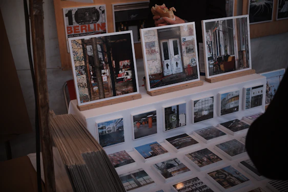 A display of art prints and photographs featuring urban and graffiti-covered scenes is set up on a table. Larger framed images rest on wooden stands, while a series of smaller prints are laid out before them. There are also visible stacks of prints to the side. Behind the display, a person holding a sandwich is partially visible.