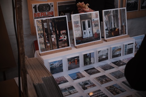 A display of art prints and photographs featuring urban and graffiti-covered scenes is set up on a table. Larger framed images rest on wooden stands, while a series of smaller prints are laid out before them. There are also visible stacks of prints to the side. Behind the display, a person holding a sandwich is partially visible.