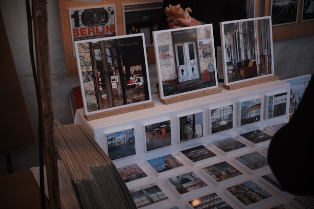A display of art prints and photographs featuring urban and graffiti-covered scenes is set up on a table. Larger framed images rest on wooden stands, while a series of smaller prints are laid out before them. There are also visible stacks of prints to the side. Behind the display, a person holding a sandwich is partially visible.
