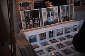 A display of art prints and photographs featuring urban and graffiti-covered scenes is set up on a table. Larger framed images rest on wooden stands, while a series of smaller prints are laid out before them. There are also visible stacks of prints to the side. Behind the display, a person holding a sandwich is partially visible.