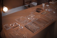 An assortment of jewelry pieces, including necklaces, rings, and earrings, is displayed neatly on a tabletop. The jewelry is organized in transparent trays, showcasing a variety of designs and sizes. The tabletop is covered with a beige cloth, and there is soft lighting illuminating the setup. Additional elements like a cup and some stationery items are present in the background.