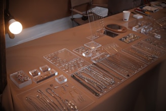 An assortment of jewelry pieces, including necklaces, rings, and earrings, is displayed neatly on a tabletop. The jewelry is organized in transparent trays, showcasing a variety of designs and sizes. The tabletop is covered with a beige cloth, and there is soft lighting illuminating the setup. Additional elements like a cup and some stationery items are present in the background.