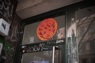 A glass door with graffiti and a colorful artistic sticker depicting a smiling face. The door is surrounded by a dark green metal frame and brick walls with various tags and stickers.