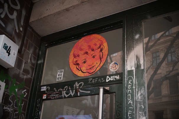 A glass door with graffiti and a colorful artistic sticker depicting a smiling face. The door is surrounded by a dark green metal frame and brick walls with various tags and stickers.