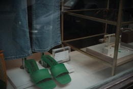 A display window featuring a pair of green slide sandals with the brand name 'BIANCO' and a pair of blue denim jeans hanging above them. To the right, various jewelry items, including necklaces and earrings, are arranged on white stands. A small price sign is visible next to the sandals.