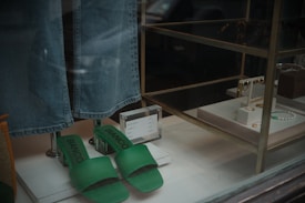 A display window featuring a pair of green slide sandals with the brand name 'BIANCO' and a pair of blue denim jeans hanging above them. To the right, various jewelry items, including necklaces and earrings, are arranged on white stands. A small price sign is visible next to the sandals.