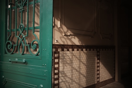 A partially open green door with intricate metalwork stands in front of a dimly lit interior space. The walls feature decorative molding and are covered with cream and dark brown tiles. Sunlight casts geometric shadows onto the wall, creating a play of light and darkness.