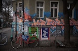An urban street scene featuring a vibrant, graffiti-covered building exterior with bold, stylized letters and cityscape motifs. In front, bicycles are parked beside a pole laden with posters, including an advertisement for a cultural event. Trees with bare branches line the sidewalk, and the surroundings convey a mix of art and everyday urban life.