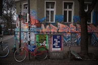 An urban street scene featuring a vibrant, graffiti-covered building exterior with bold, stylized letters and cityscape motifs. In front, bicycles are parked beside a pole laden with posters, including an advertisement for a cultural event. Trees with bare branches line the sidewalk, and the surroundings convey a mix of art and everyday urban life.